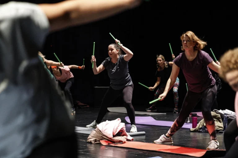 Mehrere Personen führen eine Bewegungsübung mit Stäben auf einer Bühne aus