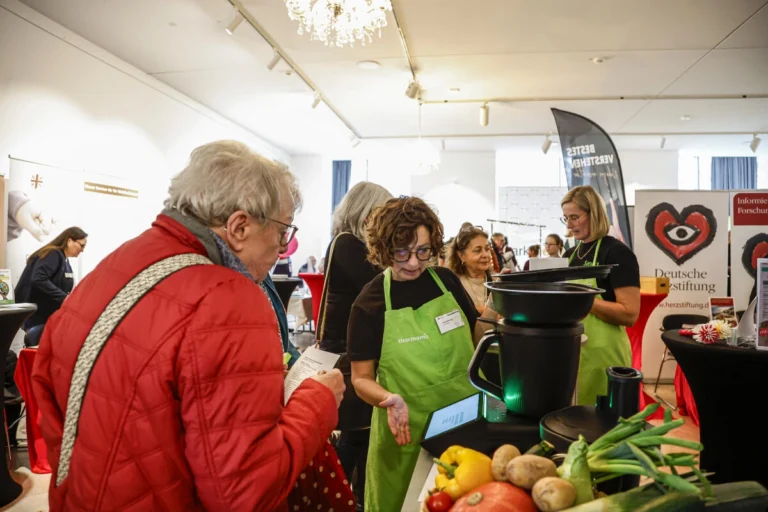 Besucherinnen und Besucher informieren sich an einem Stand mit Obst und Gemüse über ein Kochgerät