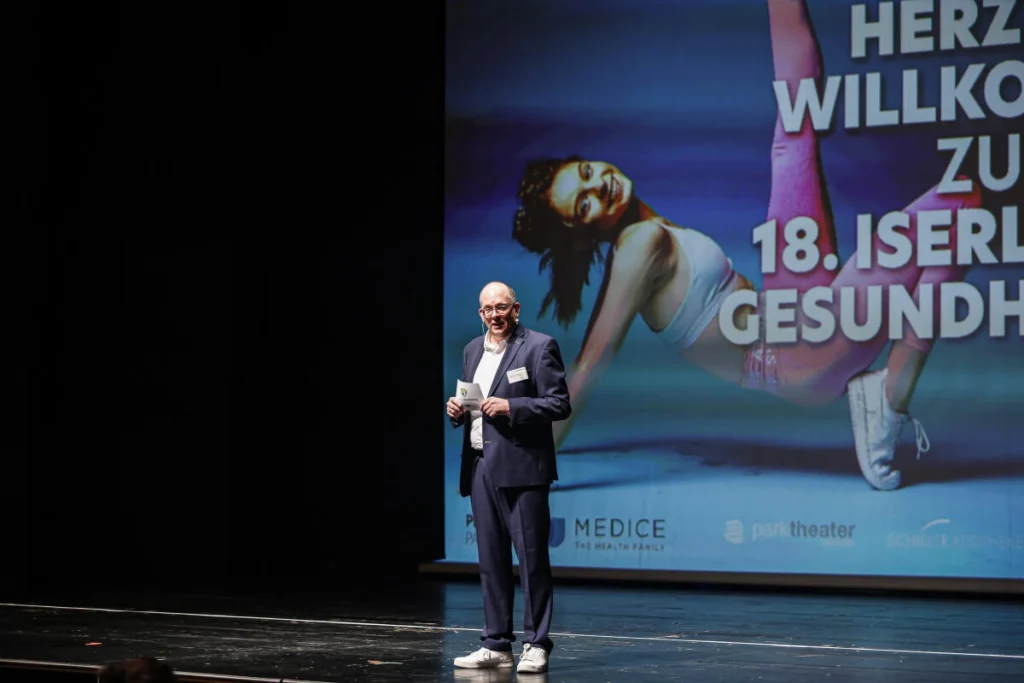 Moderator steht auf einer Bühne vor einer Leinwand mit Plakat zu einer Gesundheitsveranstaltung